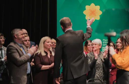 Adam Price holding a Plaid Cymru flower sign on stage while supporters applaud.