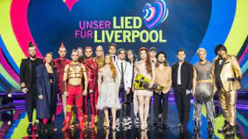 Performers standing on a stage beneath a sign reading “Unser Lied für Liverpool”.