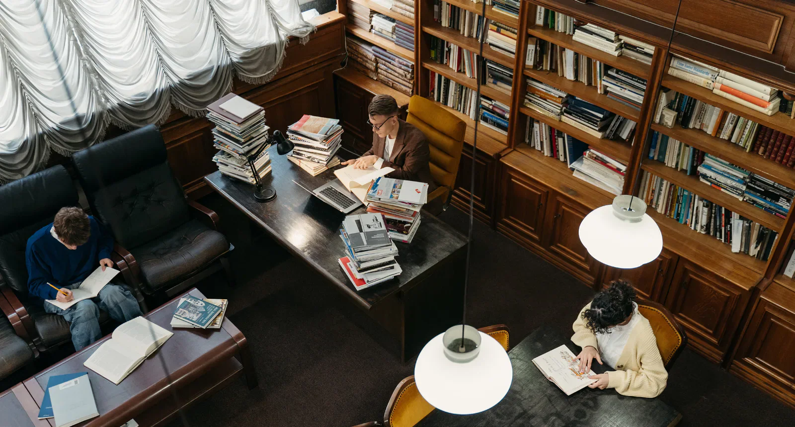 Three people read and study among bookshelves in a library.
