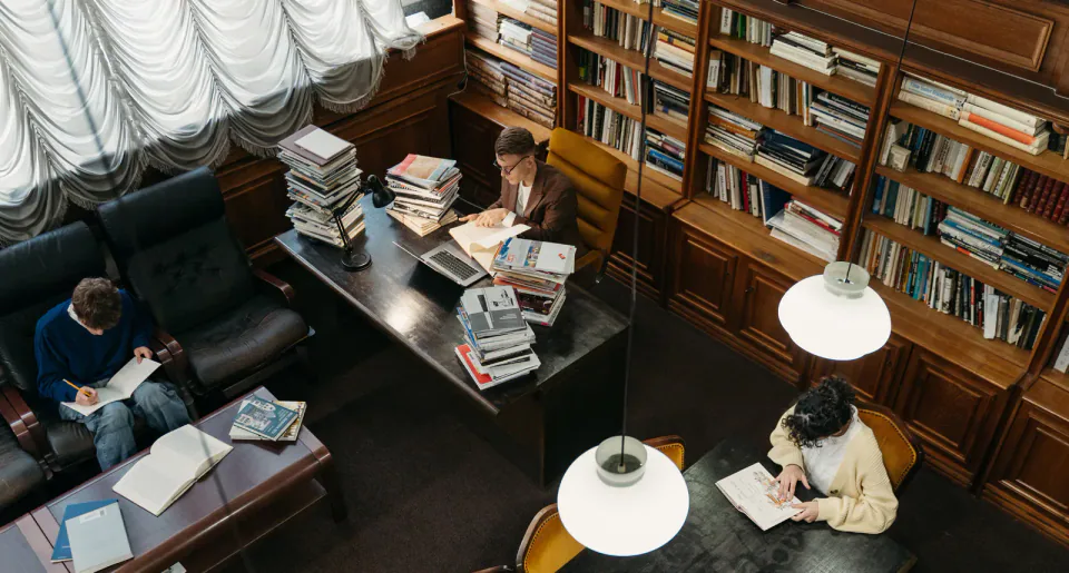 Three people read and study among bookshelves in a library.