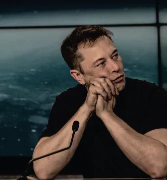 Elon Musk seated at a microphone with his chin resting on his hands