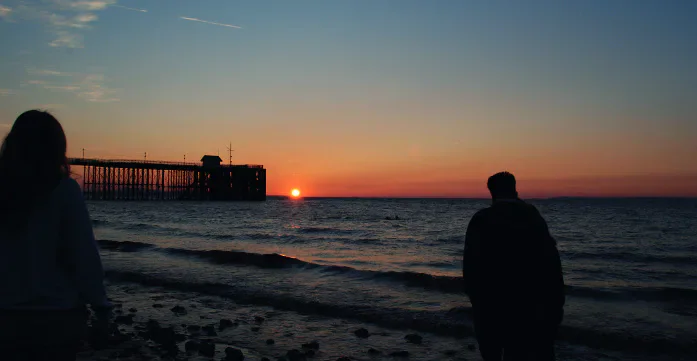 Two people silhouetted on a beach at sunset beside a pier