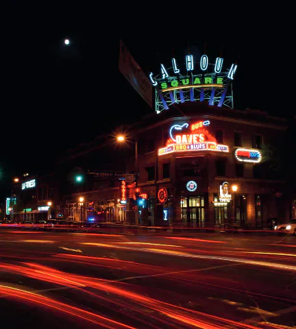 Neon signs outside a comedy club at night, with vehicle light trails in the street.
