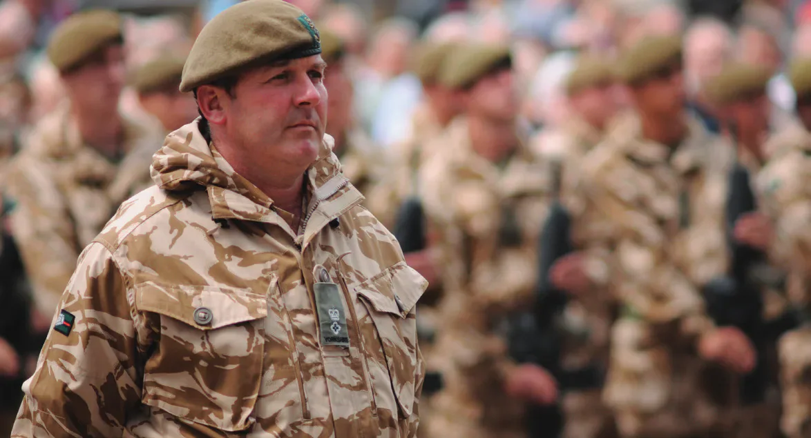 A British Army officer in uniform with soldiers behind him.