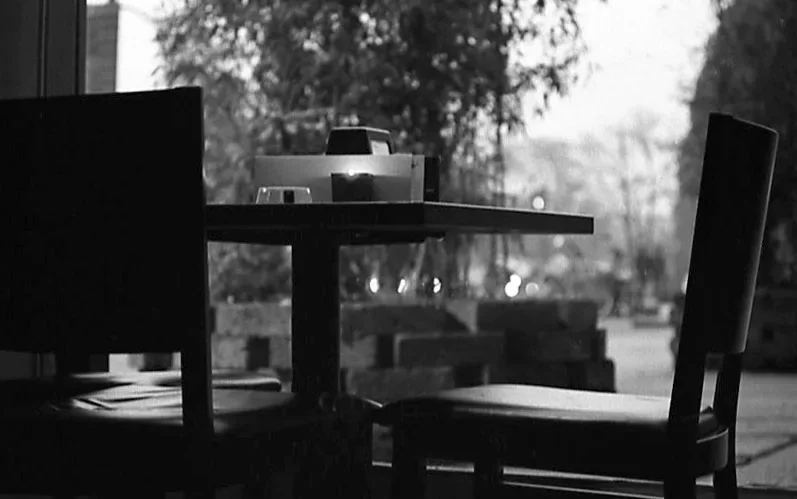 An empty café table and chairs beside a window, photographed in black and white.