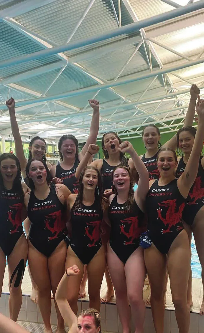 Cardiff women's water polo players in team swimsuits celebrate beside an indoor pool.