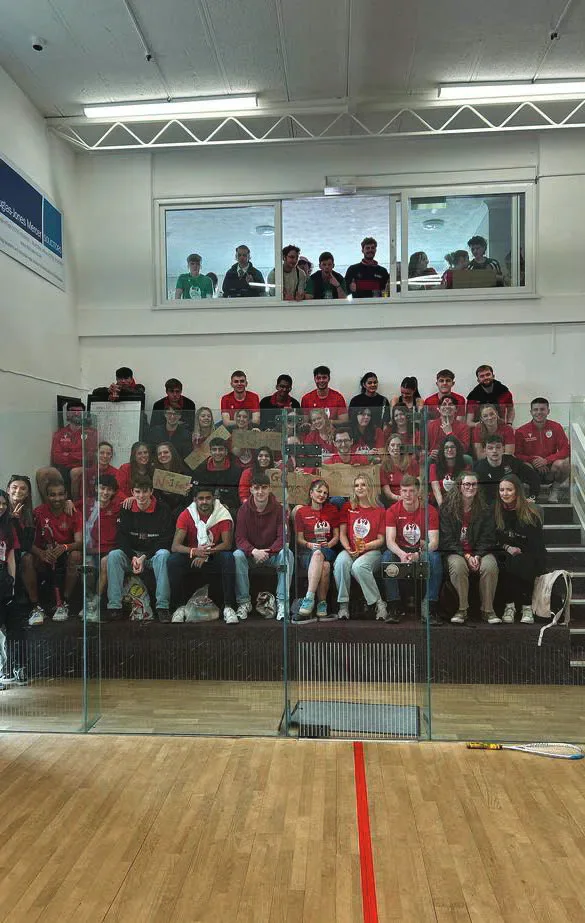 Cardiff squash players and supporters pose together behind the glass wall of an indoor squash court.