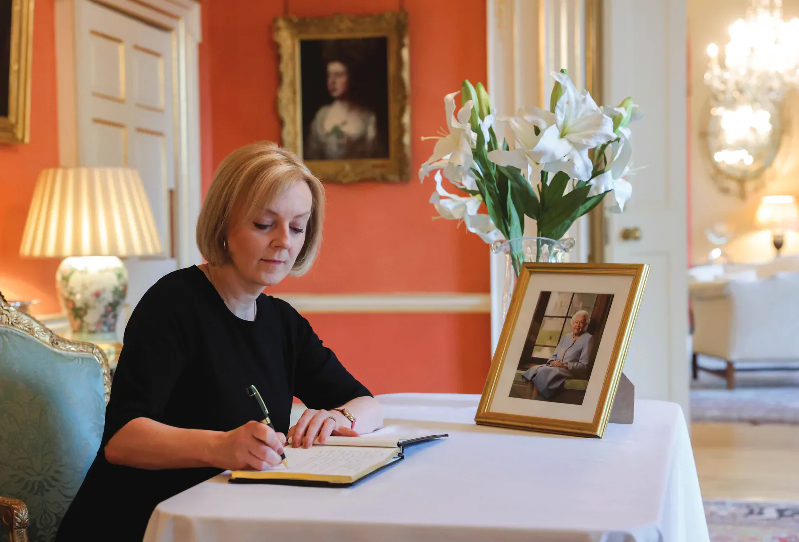 Liz Truss writing at a table beside a framed photograph of Queen Elizabeth II