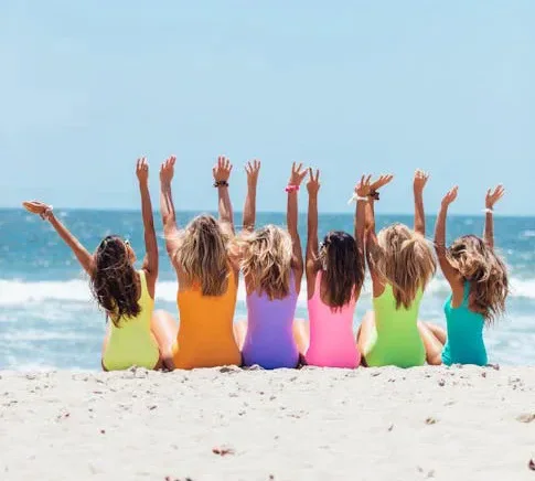 A group of girls in colourful swimsuits raising their arms on a beach.