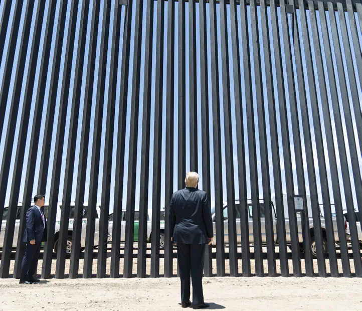 Donald Trump standing beside a tall border barrier.