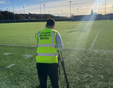 Student camera operator beside a football pitch.