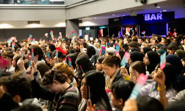 Students raising AGM voting cards in a crowded Students’ Union venue.