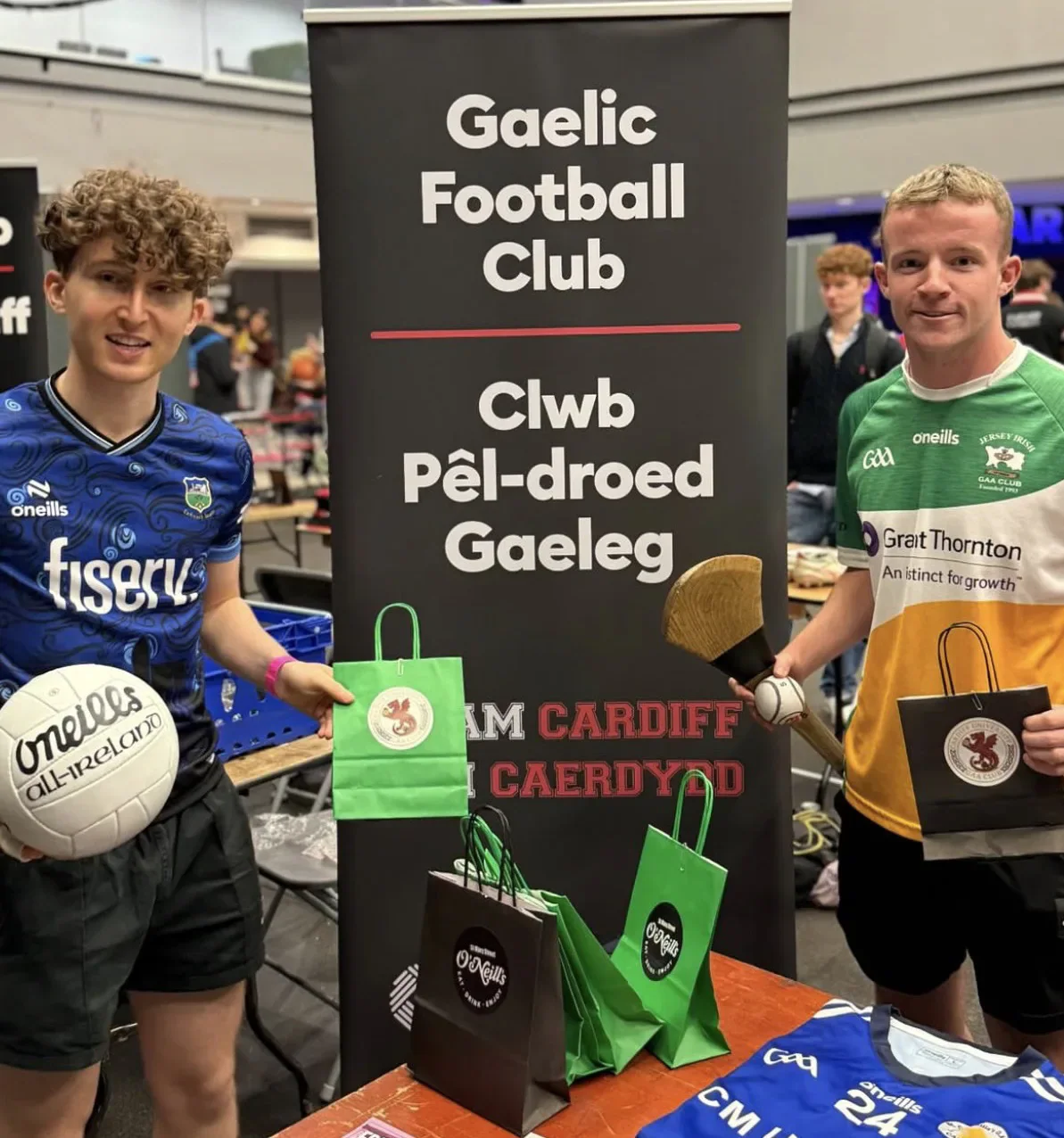 Two Cardiff Gaelic Football Club representatives beside their bilingual club banner and equipment at a fair.