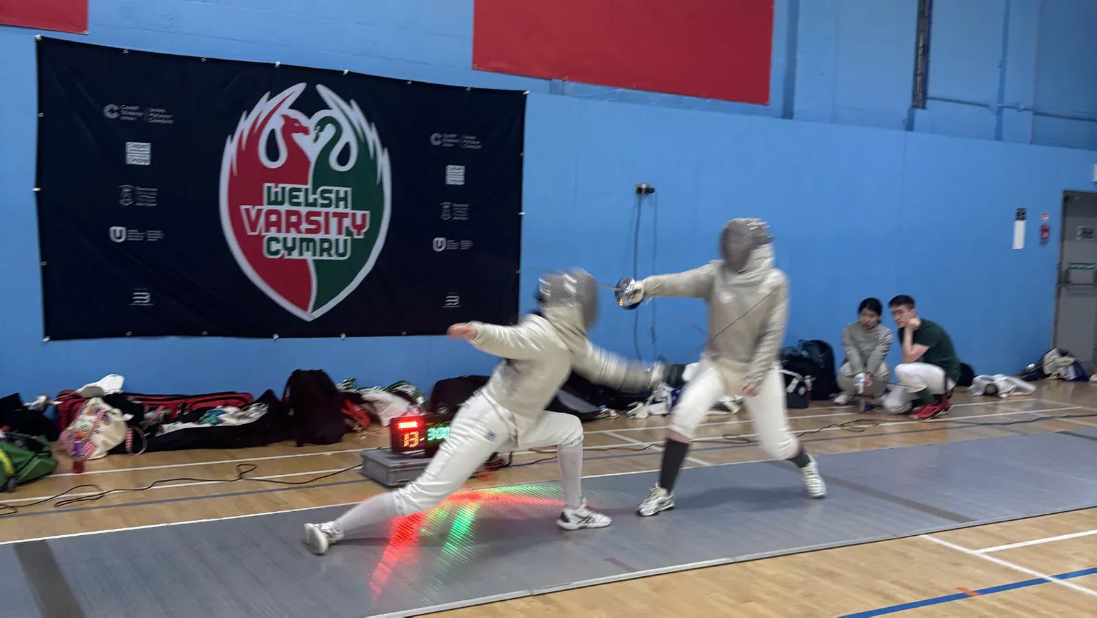 Two fencers exchange lunges beneath a Welsh Varsity banner.