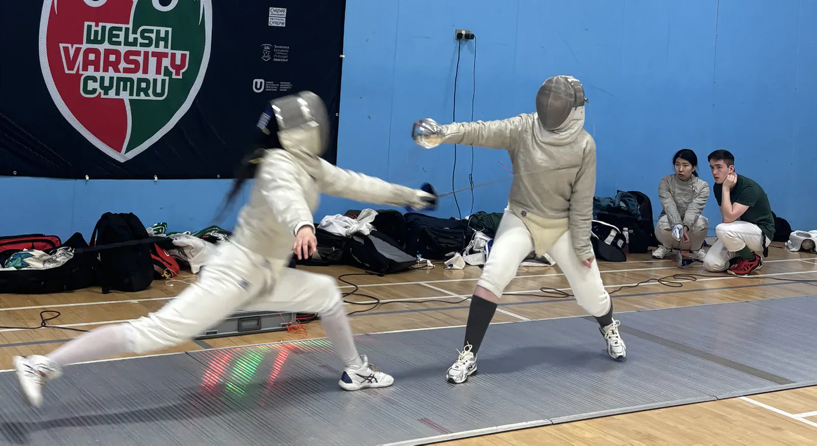 A fencer lunges forward while an opponent guards on the fencing strip.
