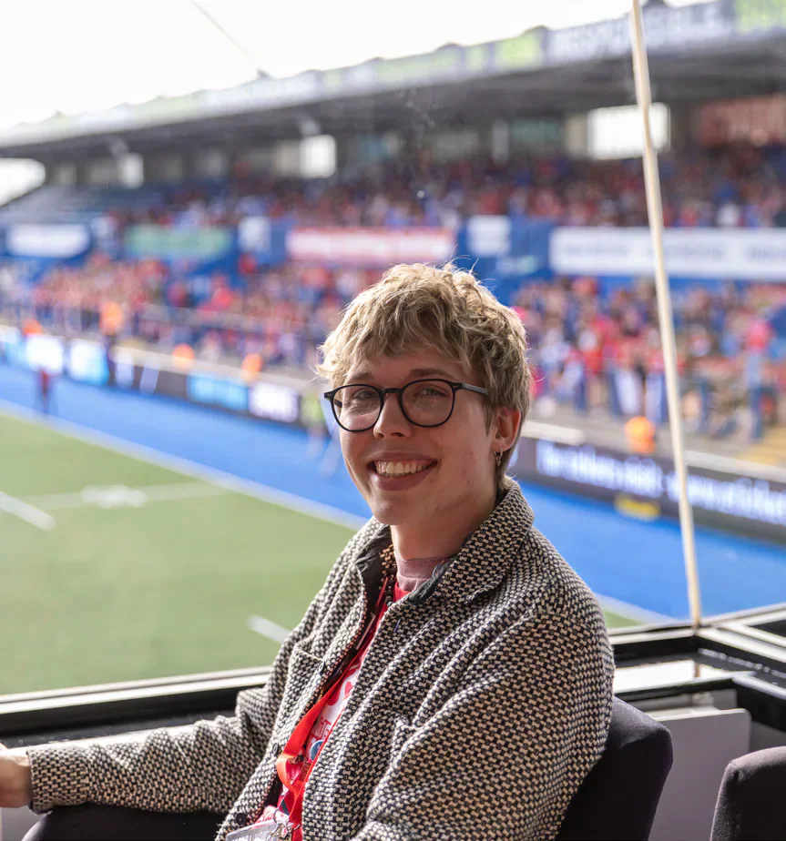 Tandy sits beside a rugby pitch with spectators in the stands behind.