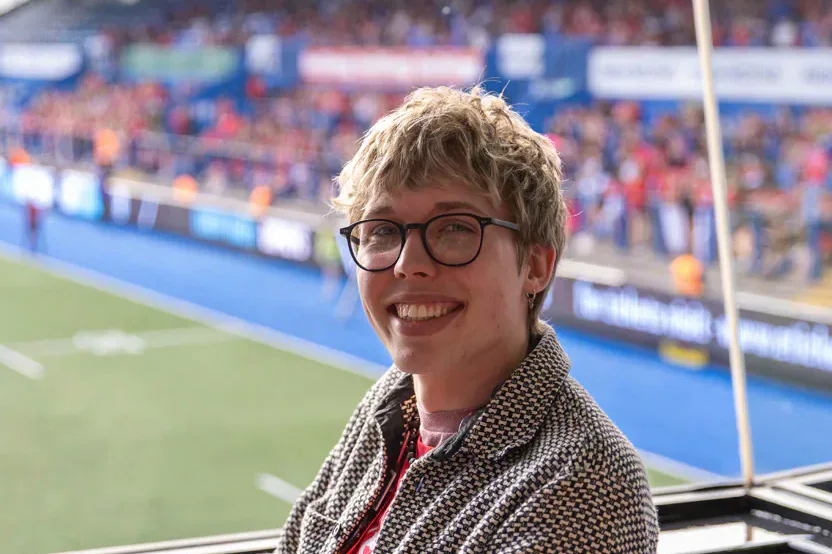 Tandy sits beside a rugby pitch with spectators in the stands behind.