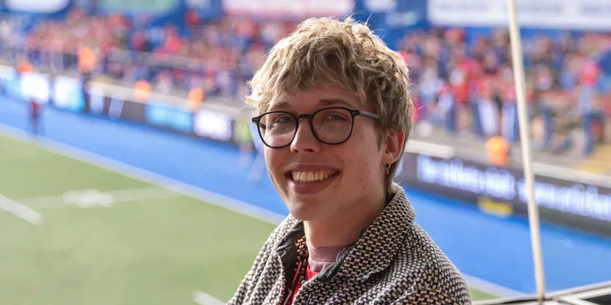 Tandy sits beside a rugby pitch with spectators in the stands behind.