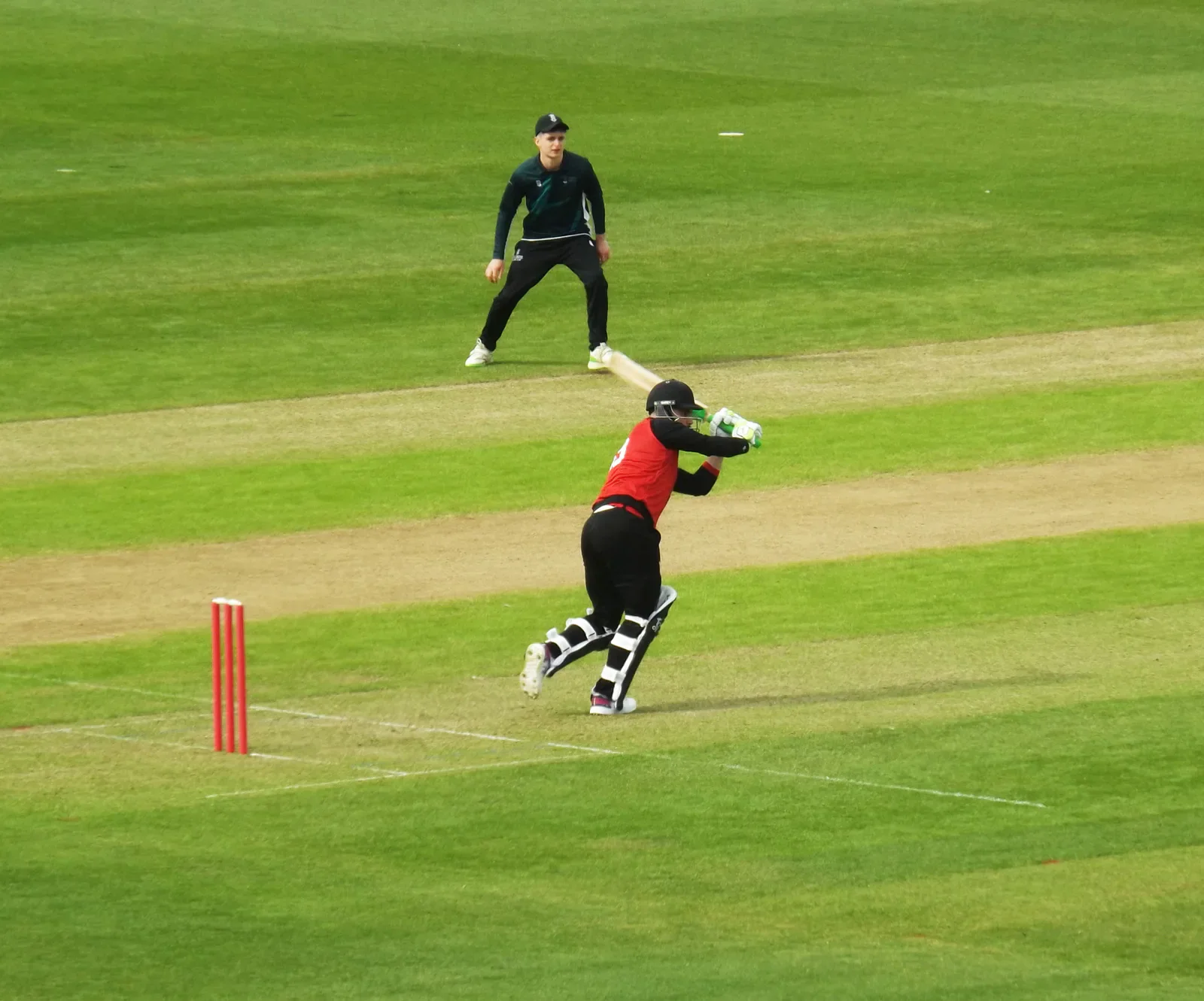 A Cardiff batter plays a shot with a Swansea fielder behind.
