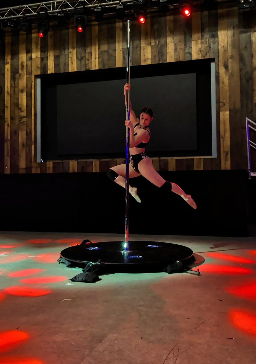 An aerial fitness competitor performs a pose on the pole under stage lighting.
