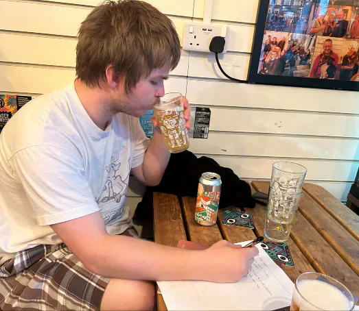 A person drinks from a pint glass while holding a pen over a quiz sheet at a pub table.