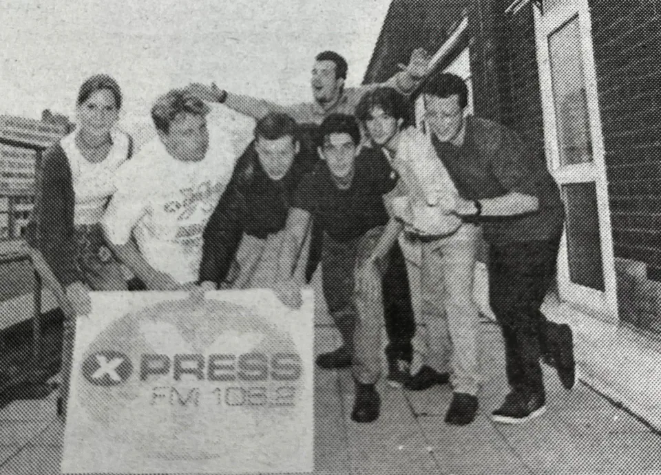 An archival black-and-white group photograph of the Xpress Radio team beside an Xpress banner.