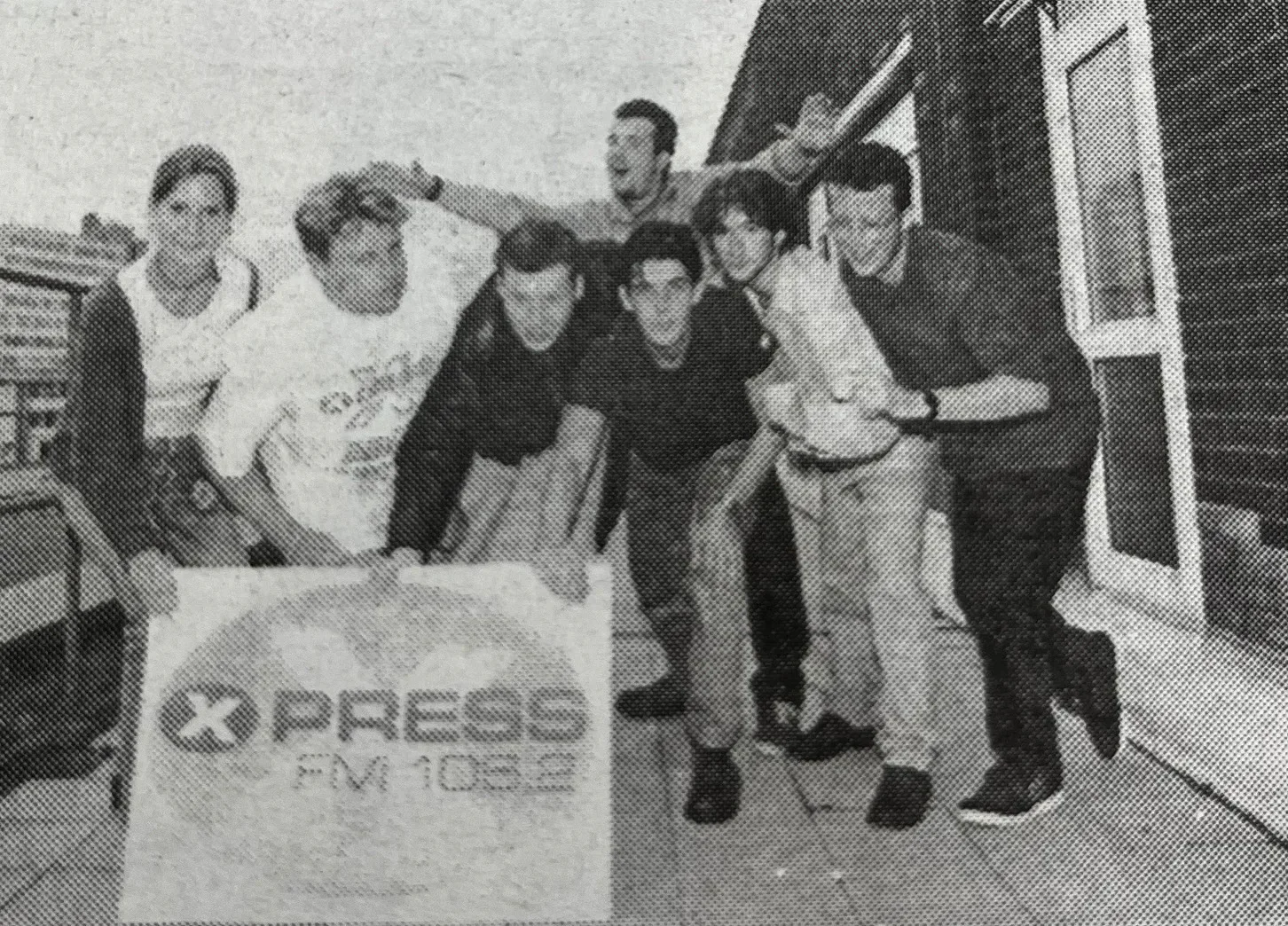 An archival black-and-white group photograph of the Xpress Radio team beside an Xpress banner.