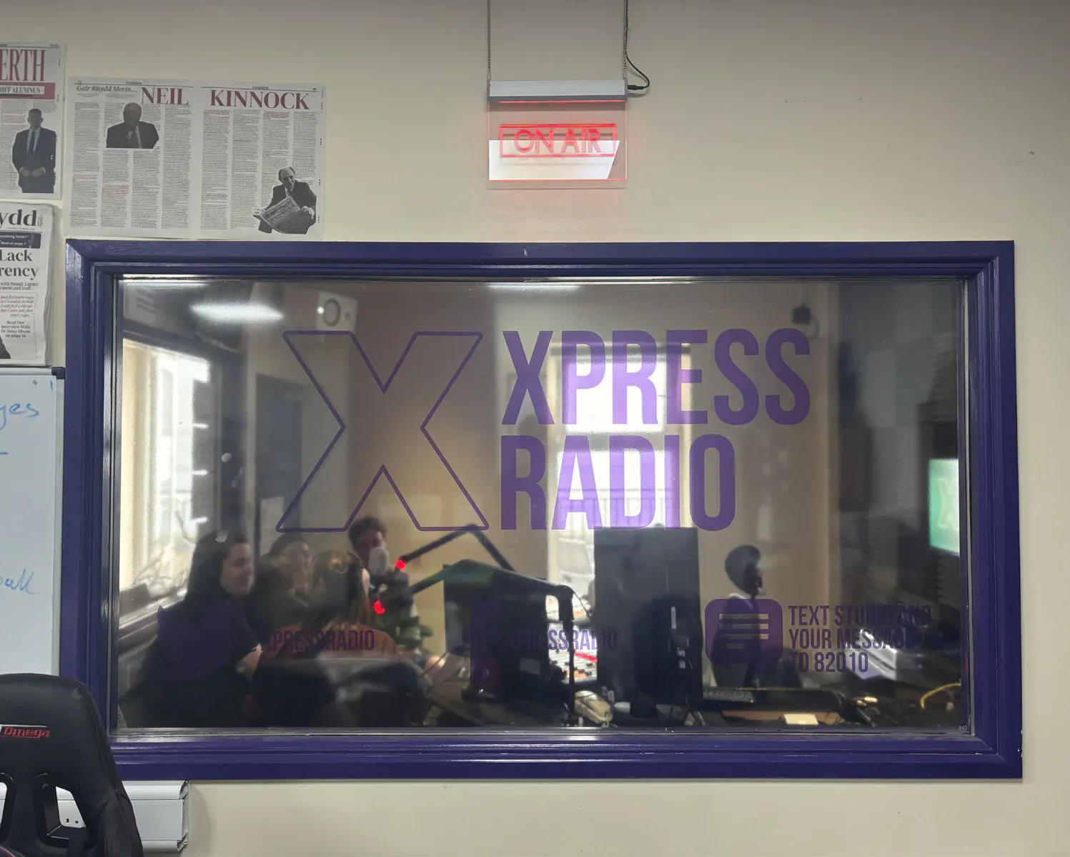 The Xpress Radio studio seen through a branded window beneath an illuminated on-air sign.