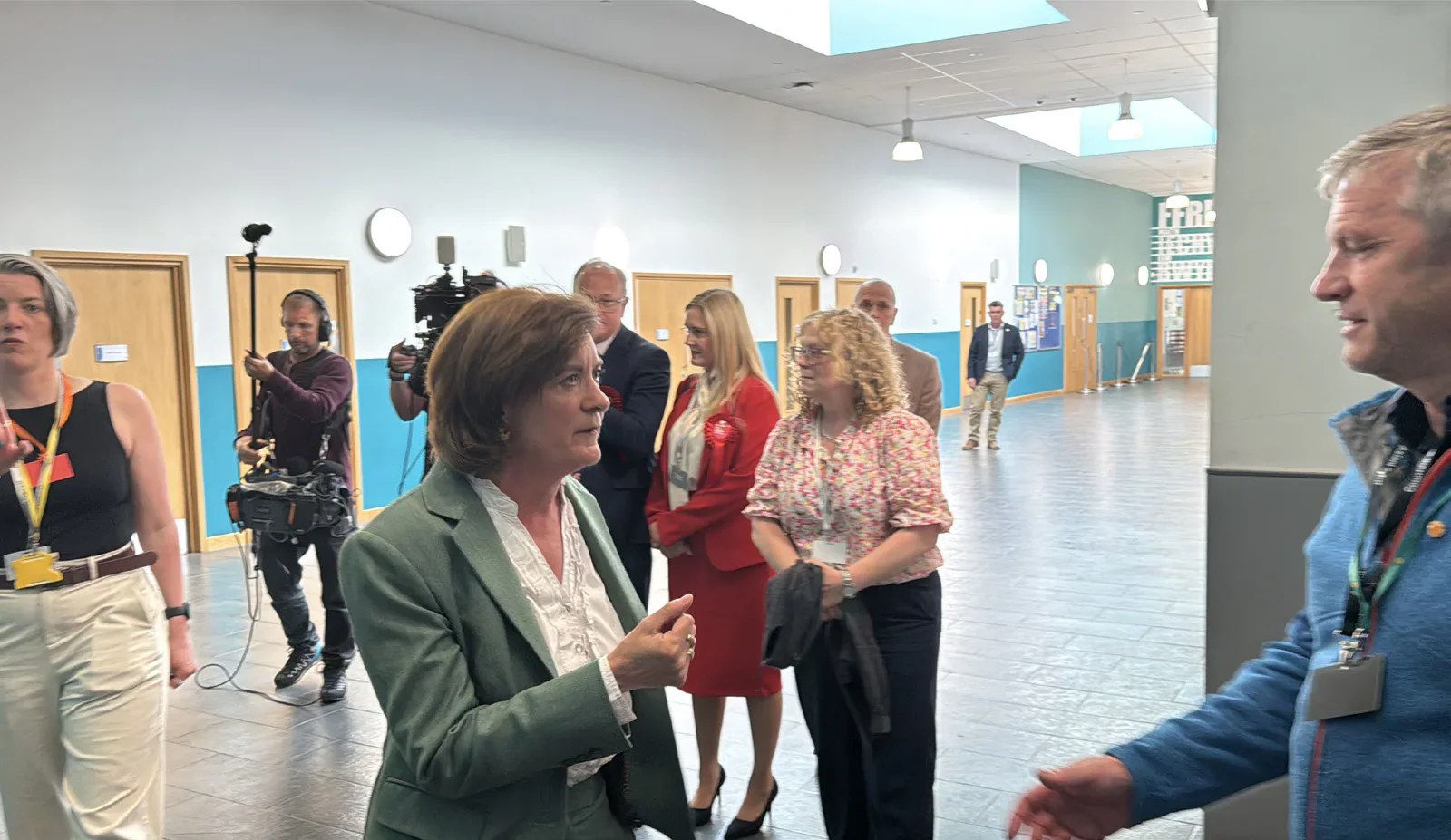 A woman in a green jacket speaking to a man inside a hall, with journalists and other people behind them.
