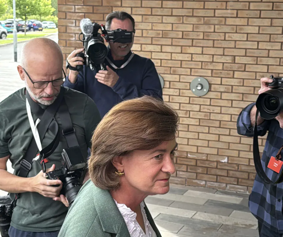 A woman in a green jacket surrounded by journalists and camera operators outside a brick building.