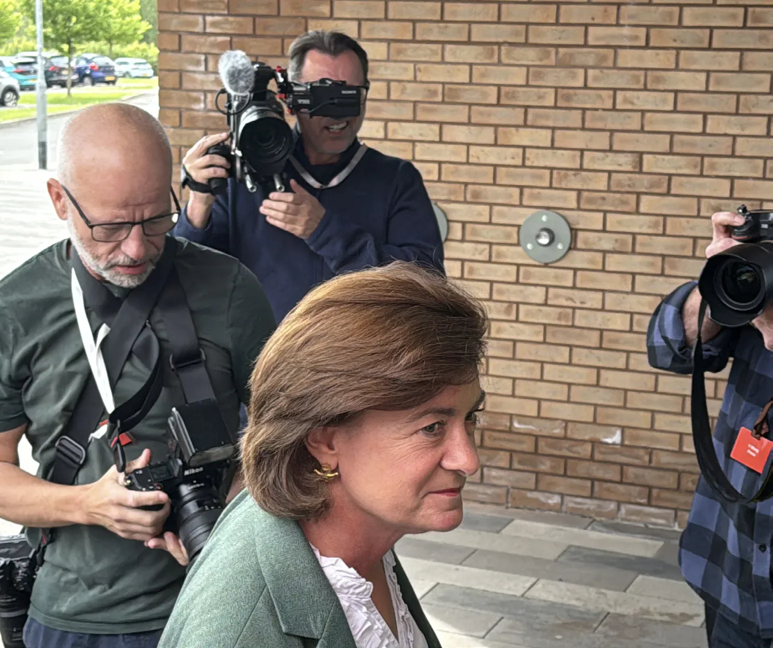 A woman in a green jacket surrounded by journalists and camera operators outside a brick building.