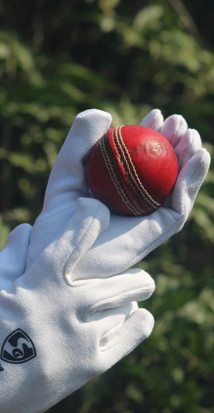 A white cricket glove holding a red cricket ball against a blurred green background.